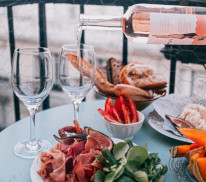 Aus einer Flasche mit Rosé-Wein wird in ein Glas eingefüllt, vor den Gläsern steht ein Teller mit Speisen