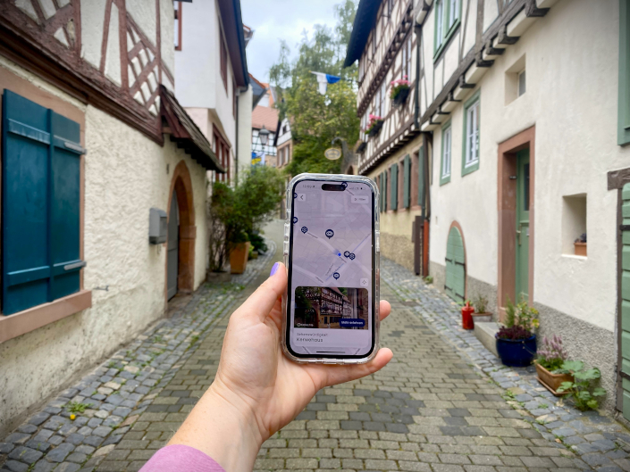 eine Hand hält ein Handy hoch auf dem ein Stadtplan mit eingezeichneten Seehnswürdigkeiten zu sehen ist, im Hintergrund sieht man die Atlstadt von Weinehim